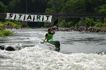 「START」と書かれた旗を後ろに河川をカヌーで進む男性の写真