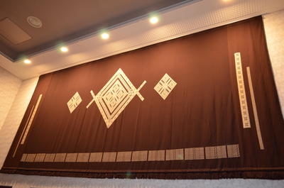 中央に菱形の模様が書かれた茶色の大きな緞帳が、壁に飾られている写真