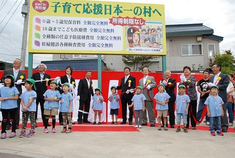 「子育て応援日本一の村へ」と書かれた看板の下でスモックを着た子供たちとスーツ姿の大人が並んで上を見ている写真